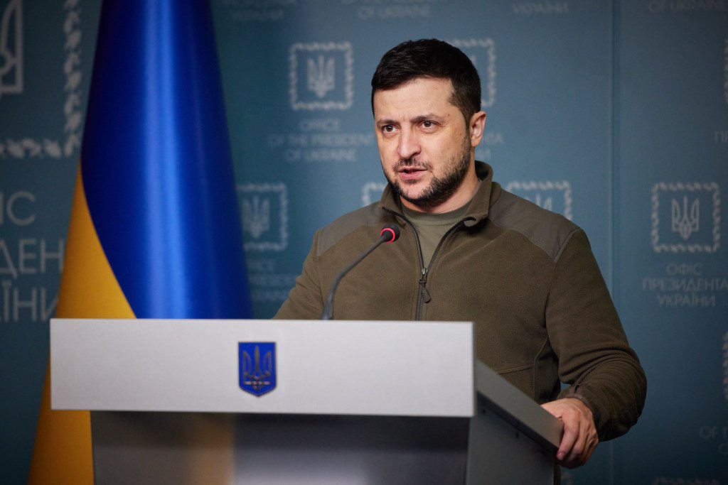 Volodymyr Zelensky speaking at a podium bearing the Ukrainian trident and flag